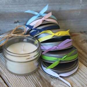 May include: A stack of small glass jars with metal lids, each tied with a different colored ribbon. The jars are filled with white candle wax and have a single wick.
