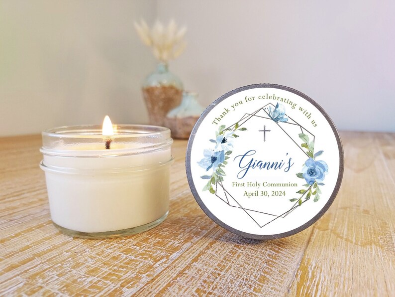 May include: A white candle in a clear glass jar with a blue and green floral design label. The label says "Thank you for celebrating with us" and "Gianni's First Holy Communion April 30, 2024".