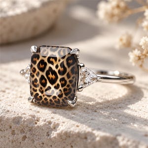 May include: A silver-toned ring featuring a large, rectangular-cut center stone with a brown and black leopard print pattern. The center stone is held by four prongs and is flanked on each side by a smaller, clear, triangular-cut side stone.