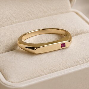 May include: A gold band ring featuring a flat, rectangular top surface with a single, small, square-cut red gemstone set into one side. The ring is displayed inside a cream-coloured jewellery box with the text KYLLIE JEWELRY printed in gold.