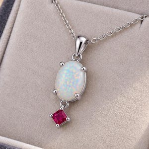 May include: A silver-toned necklace featuring an oval-shaped white opal pendant with a small, square-cut red gemstone dangling beneath it. The pendant is suspended from a delicate silver chain and presented inside a grey velvet-lined jewelry box.