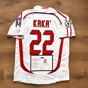 Puede incluir: Una camiseta de fútbol blanca con detalles en rojo, que muestra el nombre KAKA' y el número 22 en rojo en la parte posterior. La camiseta incluye un parche de la Champions League en la manga y un certificado de autenticidad rectangular en la parte baja.