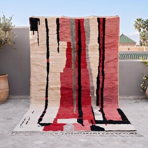 May include: A rectangular area rug featuring a cream-colored base with bold, vertical abstract stripes in shades of red, black, and gray. The design includes irregular, hand-drawn style lines and a fringed edge along the bottom of the textile.