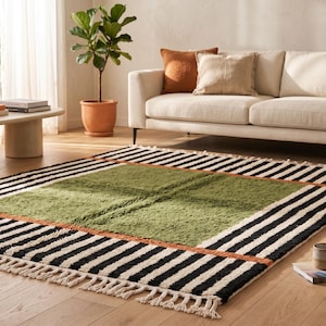 May include: A rectangular area rug featuring a large central olive green square bordered by a thin orange line and a wide frame of black and white stripes. The rug has a textured pile and includes off-white fringe along the two shorter ends.