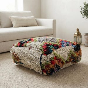 May include: A square floor pouf featuring a shaggy, textured surface with a multi-colored patchwork design. The textile is composed of cream, red, blue, green, and black fibers woven into a dense, knotted pile that creates a thick, cushioned appearance.