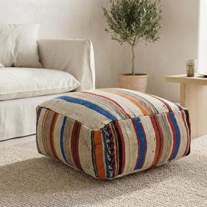 Op de afbeelding: Een vierkante vloerpoef met een geweven textielhoes met verticale strepen in tinten beige, blauw, oranje en donkerrood. De stof heeft een getextureerd, natuurlijk uiterlijk met zichtbare weefpatronen over de bovenkant en zijkanten van het kussen.