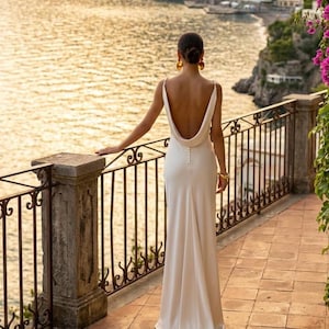 May include: A floor-length, off-white gown featuring a deep cowl back, thin shoulder straps, and a vertical row of small, round buttons running down the center of the back. The fabric is smooth and drapes toward the floor in a straight silhouette.