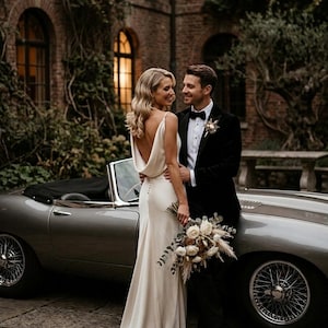 May include: A floor-length cream-colored gown with a cowl back and button detailing, paired with a black velvet tuxedo, a white dress shirt, a black bow tie, and a bouquet of white roses and dried foliage, standing beside a vintage silver car.