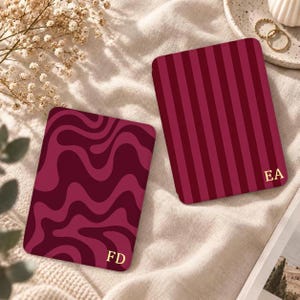 May include: Two rectangular luggage tags in shades of burgundy and dark red. One tag features a wavy, abstract pattern with the gold initials FD, while the other displays vertical stripes and the gold initials EA in the bottom corner.