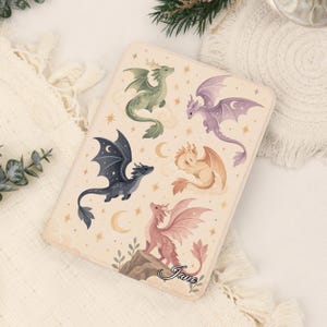 May include: A rectangular e-reader case featuring a cream background with illustrations of four dragons in green, purple, gold, and pink. The design includes stars, crescent moons, and clouds, with the name Jane written in cursive script at the bottom.