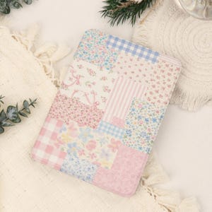 May include: A rectangular protective case featuring a patchwork design of various floral patterns, pink and blue gingham checks, and vertical stripes in soft pastel shades of pink, blue, and white, with rounded corners and a smooth, flat surface.