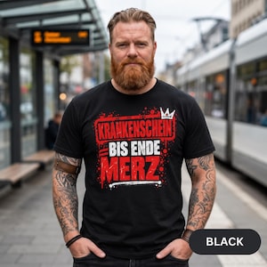 Könnte beinhalten: Ein schwarzes Kurzarm-T-Shirt mit einem grafischen Aufdruck, der den Text KRANKENSCHEIN BIS ENDE MERZ in roten und weißen Blockbuchstaben zeigt, ergänzt durch ein kleines weißes Kronensymbol und einen Farbspritzer-Effekt.
