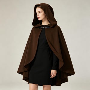 May include: A dark brown hooded cape featuring a smooth, solid-colored fabric with a flared, flowing silhouette. The garment is secured at the neckline with a decorative black metal clasp, providing a structured closure over a simple black dress.