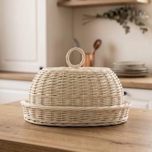 May include: A light beige woven wicker food cover with a dome shape and a circular handle on top. The cover features a flat, circular base rim and a tightly woven lattice pattern throughout, providing a natural texture and a neutral color palette.