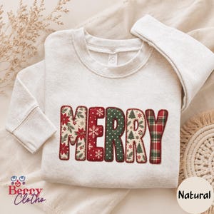 May include: A cream-coloured crewneck sweatshirt featuring the word MERRY appliqued in large, block letters. Each letter displays a different holiday-themed pattern, including poinsettias, snowflakes, polka dots, pine trees, and red and green plaid.