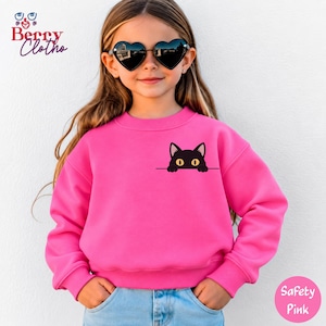 May include: A bright pink long-sleeve sweatshirt featuring a graphic of a black cat with yellow eyes peeking over a horizontal line on the chest. The sweatshirt has a crew neckline and ribbed cuffs, paired with light blue denim jeans.