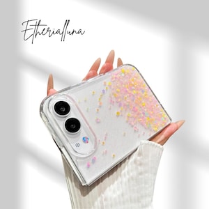 May include: A clear phone case for a foldable smartphone featuring a white glittery base with scattered pastel-colored confetti pieces. The case includes a cutout for a dual-camera system and a flash. The text Etherialuna is visible above.
