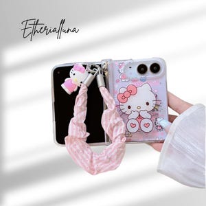 Puede incluir: Una funda de teléfono plegable transparente con un gráfico de Hello Kitty, un pequeño dije de Hello Kitty y una correa de muñeca de tela a rayas rosas y blancas. El texto Etherialuna es visible en la esquina superior izquierda.