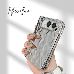 Peut inclure: Une coque de téléphone argentée et réfléchissante avec une texture ondulée semblable à du liquide et un bord transparent. La coque présente une découpe pour un système de caméra à double objectif. Le texte "Etherialfuna" est écrit en noir.