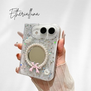 May include: A clear phone case with silver glitter and sequins, featuring a circular mirror framed by white faux pearls and a small pink bow accent. The text Etherialuna is printed in black script above the case, which has two camera cutouts.