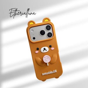 May include: A brown silicone phone case featuring a raised Rilakkuma bear character holding a pink and white swirl lollipop. The case has bear ears at the top, a camera cutout, and the text Rilakkuma and Etherialuna printed on the surface.