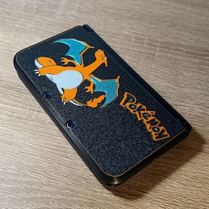 Könnte beinhalten: Eine schwarze tragbare Spielkonsole mit texturierter Oberfläche, die mit einer Grafik des orange-blauen drachenartigen Charakters Glurak und dem orangefarbenen Pokémon-Logo auf der Oberseite des geschlossenen Geräts versehen ist.