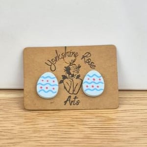 May include: Easter egg stud earrings in light blue with white and red patterns, presented on a brown card. The card has the text "Yorkshire Rose Arts". The earrings are on a wooden surface. The background is a light brown wooden surface.