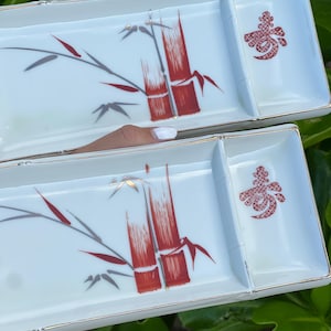 May include: Two rectangular white ceramic serving dishes with a gold-trimmed edge. Each dish features a painted design of red and grey bamboo stalks and a red decorative symbol on the right side. The dishes are divided into two sections.