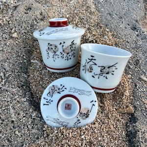 Può includere: Un set da tè in porcellana bianca composto da una ciotola con coperchio e una tazza abbinata. Entrambi i pezzi presentano scritte calligrafiche nere, una texture a coste verticali e una fascia rosso scuro alla base e sul bordo.