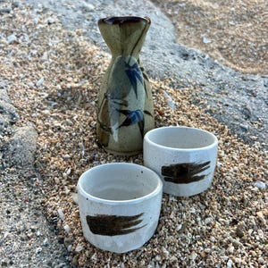 Può includere: Un set da sake in ceramica composto da una bottiglia alta dal collo stretto con finitura beige maculata e motivi di foglie di bambù blu scuro, e due piccole tazze bianche maculate abbinate con un tratto di pennello marrone scuro.