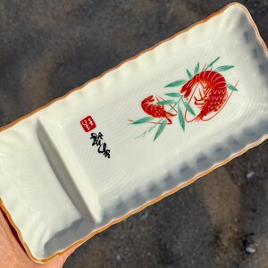 May include: A rectangular white ceramic serving tray with a scalloped edge and a thin orange rim. The surface features a graphic of two red shrimp among green bamboo leaves, a black calligraphic inscription, and a small red square seal mark.