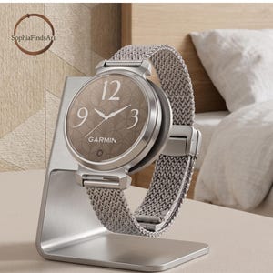 May include: A silver Garmin smartwatch with a round face, a brown dial featuring a fan-like pattern, white numerals at the 3, 9, and 12 positions, and a silver mesh metal band, displayed on a silver-toned metal stand.