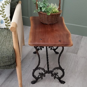 May include: A small, rustic side table with a dark wood top and ornate black metal legs. A woven basket filled with greenery sits on the table. The table is next to a green upholstered chair.