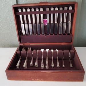 May include: A wooden storage chest containing a set of silver-toned flatware. The top section holds twelve knives arranged vertically, while the bottom section contains various forks and spoons organized in individual slots with dark brown lining.