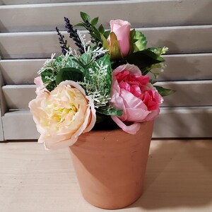 May include: A terracotta pot filled with an arrangement of artificial flowers. The arrangement includes pink and peach-colored peonies, lavender sprigs, and green foliage. The pot sits on a light-colored wooden surface.