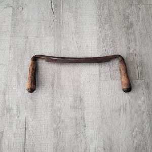 May include: A drawknife featuring a long, slightly curved metal blade with a dark, weathered finish. The tool includes two cylindrical wooden handles, one on each end, showing signs of age and wear with a natural, light brown wood grain texture.