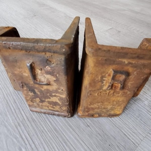 May include: Two weathered, rusty metal objects with angled sides and a rectangular base. One has an "L" and the other has an "A" stamped on the side. The objects are likely industrial or architectural salvage.