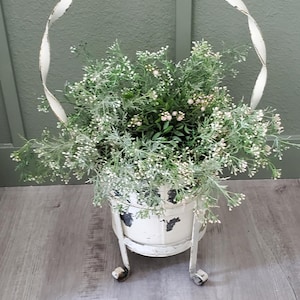 May include: A distressed white metal planter with a twisted handle, filled with greenery and small white flowers. The planter sits on small wheels and has a weathered appearance, suggesting a vintage or farmhouse style. The overall aesthetic is rustic and charming.