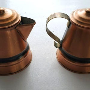 May include: Two copper-colored metal coffee pots with brass handles and lids. The pots have a rounded base and a black band around the middle. One pot has a spout. The pots are likely vintage or antique.