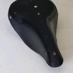 May include: A black bicycle saddle with a smooth, curved surface and a tapered nose. The seat features two circular holes near the rear and small metallic rivets along the sides and front, showing a classic design with a glossy finish.
