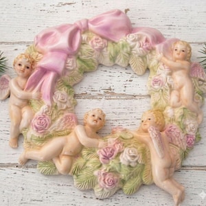 May include: A circular decorative wall plaque featuring three cherubs with wings nestled among sculpted roses and foliage. A large, prominent pink bow is positioned at the top center of the wreath. The entire piece is finished in soft pastel tones of pink, cream, and pale green, creating a detailed floral arrangement with angelic figures.