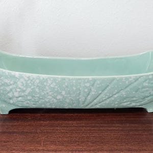 May include: A rectangular, light-green ceramic planter with a speckled texture and leaf design. The planter sits on four small feet and is displayed on a dark wooden surface. Lavender sprigs are on the left.
