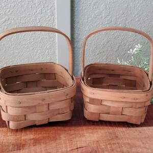 May include: Two square-shaped woven wooden baskets with light brown finish and fixed arched handles. Each basket features a horizontal and vertical slat construction with visible metal fasteners along the top rim, providing a sturdy structure.