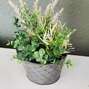 May include: A potted arrangement featuring a variety of artificial greenery and tall, pale cream-colored stalks. The plants are contained within a round, gray metal bucket that has a textured, diamond-patterned surface embossed on its exterior.