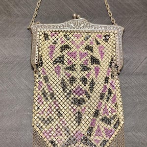 Può includere: Una borsa in maglia metallica con un motivo geometrico color crema, nero e viola. La borsa presenta una cornice in metallo argentato con un design in rilievo, un sottile manico a catena e un bordo inferiore a punta con frange metalliche.
