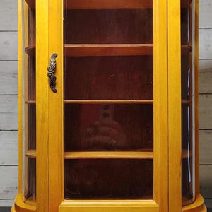 Pode incluir: Uma vitrine alta de madeira cor de mel com uma porta de vidro curva e um puxador de metal decorativo. O móvel tem um topo recortado, quatro prateleiras internas, um painel traseiro escuro e assenta sobre quatro pernas curvas.