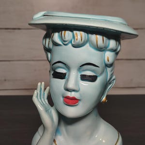 Könnte beinhalten: Eine hellblaue Keramik-Kopfvase, die eine Person mit einem breitkrempigen Hut darstellt. Die Figur hat geschlossene Augen mit dicken schwarzen Wimpern, rote Lippen und goldfarbene Akzente an Haar und Ausschnitt. Eine Hand berührt die Wange.