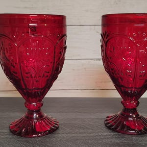 May include: A pair of deep red glass goblets featuring a raised, intricate embossed pattern on the bowl and a wide, fluted base. The stem of each glass is short and thick, connecting the textured bowl to the flared, scalloped foot.
