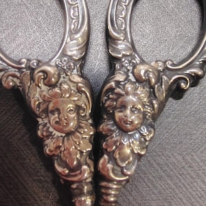 May include: A pair of silver-toned metal scissors featuring ornate, embossed handles. Each handle is decorated with a detailed relief of a cherubic face surrounded by intricate floral and scrollwork patterns, extending into circular finger loops.