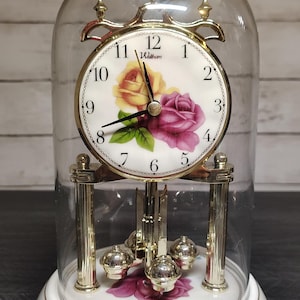 May include: A Waltham anniversary clock featuring a white face with black numerals and a floral design of yellow and purple roses. The clock is encased in a glass dome, sits on a white ceramic base with floral patterns, and has gold-tone accents.